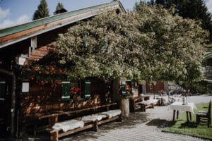 Eine rustikale Holzhütte mit grünen Fensterläden und roten Blumen, perfekt für eine bezaubernde Hochzeit. Eingebettet zwischen Bäumen spendet ein großer Baum Schatten auf einer Holzterrasse mit Bänken, Tischen und einem in der Nähe geparkten Mountainbike. Üppiges Grün und blauer Himmel umhüllen diese idyllische Kulisse für Hochzeiten.