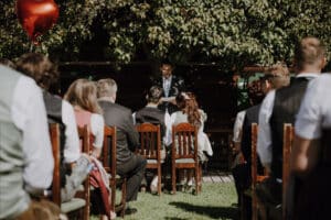 Eine intime Hochzeit findet im Freien statt, wo die Gäste auf Holzstühlen im warmen Schein der Sonne sitzen und dem Paar und dem Geistlichen unter einem großen Baum gegenüberstehen. Im linken Vordergrund hält jemand einen roten herzförmigen Ballon, der diesem wunderschön geplanten Moment in der Natur eine skurrile Note verleiht.