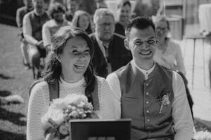 Ein glückliches Paar in traditioneller Kleidung sitzt bei einer Veranstaltung im Freien zusammen und feiert seine Hochzeit. Die Frau hält einen Blumenstrauß in der Hand. Beide lächeln, während die Leute im Hintergrund aufmerksam zuschauen. Das Schwarzweißbild fängt die zeitlose Freude ihres besonderen Tages ein.