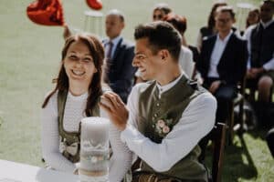 Ein Paar sitzt lachend während einer Hochzeitszeremonie im Freien, in traditioneller Kleidung und mit einer dekorierten Kerze vor ihnen. Die Gäste sitzen im Hintergrund inmitten der sonnigen, grasbewachsenen Umgebung – eine perfekte Szene für diejenigen, die sich von einer durchdachten Hochzeitsplanung inspirieren lassen.