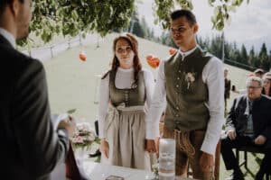 Ein Paar in traditioneller Kleidung steht draußen und hält sich während der Hochzeitszeremonie an den Händen. Im Hintergrund sind ein grasbewachsener Hügel, Bäume und sitzende Gäste zu sehen. Im Vordergrund steht ein Mann, möglicherweise ein Geistlicher, der die Hochzeitsplanung überwacht, und ist teilweise sichtbar.