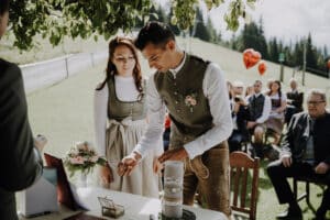 Ein Paar in traditioneller Kleidung nimmt an einer Hochzeitszeremonie im Freien teil. Der Bräutigam zündet eine Kerze an, während die Braut in seiner Nähe steht, und verkörpert damit ihre gemeinsame Reise. Die Gäste sitzen, einige halten rote herzförmige Luftballons in einer üppigen grünen Wiese unter einem Baum.