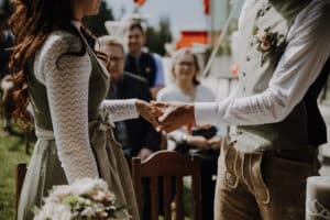 Das Paar gibt sich während seiner Hochzeit im Freien das Ja-Wort. Die Frau in einem langärmeligen Kleid mit Spitzendetails hält einen Blumenstrauß. Der Mann trägt eine Weste mit Blumendekor und eine traditionelle Hose. Die Gäste machen es sich in der Sonne gemütlich, um diesen wunderschönen Moment der Hochzeit mitzuerleben.