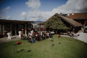 Eine bezaubernde Hochzeitszeremonie im Freien findet statt, bei der die Gäste auf einer Wiese gegenüber einem charmanten rustikalen Holzgebäude sitzen. Rote herzförmige Ballons verleihen der Szene eine stimmungsvolle Note und fangen die Freude der Hochzeit an diesem hellen, sonnigen Tag mit Wolken am Himmel ein.
