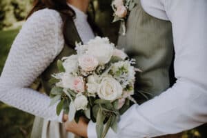 Das Paar in heller Kleidung hält einen Strauß weißer und rötlicher Rosen mit Grün in den Händen, der ihre Hochzeit symbolisiert. Sie stehen eng beieinander im Freien und fangen einen romantischen Moment ein.
