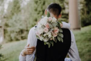 Braut und Bräutigam umarmen sich in einer üppigen Umgebung im Freien und verkörpern die Essenz einer traumhaften Hochzeit. Die Braut hält einen Strauß rosafarbener und weißer Blumen mit grünen Blättern über der Schulter des Bräutigams und trägt eine schwarze Weste. Sie stehen vor einem verschwommenen Hintergrund aus Bäumen und Gras.