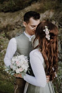In einer ruhigen, natürlichen Umgebung, die perfekt für jede Hochzeit ist, umarmt sich ein Paar zärtlich im Freien. Die Frau, geschmückt mit Blumen im Haar, hält einen Strauß weißer und rosa Rosen. Beide tragen traditionelle Kleidung: der Mann trägt eine Weste, die Frau ein strukturiertes Kleid.
