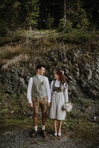 Ein Paar in traditioneller Kleidung hält sich an den Händen und blickt sich an. Es feiert seine Hochzeit inmitten eines felsigen Hügels, umgeben von viel Grün. Die Frau hält einen Blumenstrauß und trägt einen Blumenkranz, der die Essenz ihres Heiratsmoments einfängt.