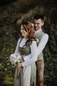 Ein Paar in traditioneller Kleidung steht in einem Wald und lächelt herzlich. Die Frau, die an der Hochzeitsplanung beteiligt ist, hält einen Blumenstrauß in der Hand und trägt eine weiße Bluse und ein grünes Kleid. Der Mann steht hinter ihr und trägt ein passendes Outfit. Sie wirken beide fröhlich und entspannt, während sie ihre Hochzeit planen.