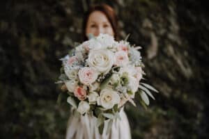 Eine Frau, die einen Strauß Rosen und Grünpflanzen mit zartrosa und weißen Blüten hält, fängt vor einem unscharfen natürlichen Hintergrund die Essenz der Hochzeitsplanung ein.