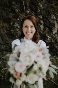 Eine lächelnde Frau mit braunem Haar steht draußen und hält einen Strauß weißer und rosa Blumen in der Hand, der die Freude einer Hochzeit verkörpert. Sie trägt ein helles Outfit, perfekt für eine Hochzeit, und der Hintergrund ist mit Felsen und Grün gestaltet.