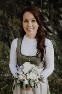 Vor der ruhigen Felskulisse hält eine Person mit langen braunen Haaren einen Strauß rosa und weißer Blumen. Sie trägt ein weißes Oberteil und ein grünes Kleid, als würde sie die Freude und Eleganz von Hochzeitsträumen und Hochzeitsplanung verkörpern.