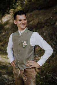 Ein Mann in traditioneller Kleidung steht im Freien, die Hände in die Hüften gestemmt, und lächelt. Er trägt ein weißes Hemd und eine grüne Weste mit einem Blumenboutonniere, dazu braune bestickte Hosen – perfekt für eine Hochzeit in einer natürlichen Umgebung aus Felsen und Gras.