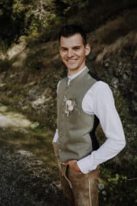Ein Mann steht lächelnd auf einem Kiesweg im Grünen. Er trägt traditionelle Hochzeitskleidung mit weißem Hemd, grüner Weste und brauner Hose. An seiner Weste ist eine kleine Blumenboutonniere angebracht.