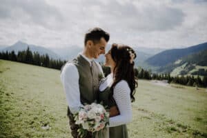 Ein Paar in traditioneller Kleidung umarmt sich im Freien auf einer Wiese mit Bergen im Hintergrund. Die Frau, die eine sorgfältige Hochzeitsplanung durchführt, hält einen Blumenstrauß und trägt Blumenschmuck für das Haar. Beide lächeln sich unter einem teilweise bewölkten Himmel an.