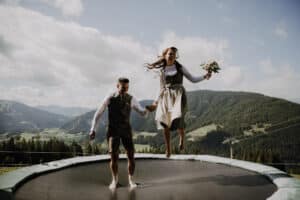 Ein Paar in traditioneller Kleidung springt fröhlich auf einem Trampolin inmitten einer atemberaubenden Berglandschaft und feiert seine Hochzeit. Die Frau hält einen Blumenstrauß und beide lächeln, mit üppigen grünen Hügeln und einem bewölkten Himmel im Hintergrund.