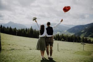 Auf einer Wiese mit Bergen in der Ferne feiert ein Paar seine Hochzeit. Die Frau hält einen Blumenstrauß, während der Mann einen roten herzförmigen Ballon umklammert. Sie umarmen sich, blicken auf die malerische Landschaft und denken über ihre gemeinsame Zukunft nach.
