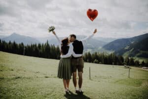 Ein Paar steht auf einem grasbewachsenen Hügel, umarmt und küsst sich, hält einen herzförmigen Ballon und einen Blumenstrauß in den Händen und feiert seine Hochzeit. Sie blicken auf eine malerische Landschaft aus Bergen und Wäldern unter einem teilweise bewölkten Himmel und träumen von der Zukunft, die sie erwartet.