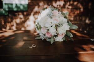 Ein Strauß weißer und rosa Blumen mit grünen Blättern steht auf einem Holztisch im gesprenkelten Sonnenlicht und bietet eine perfekte Kulisse für die Hochzeitsplanung. Neben dem Strauß liegen zwei goldene Eheringe, die das Heiratsversprechen symbolisieren.