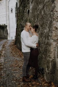 Ein Paar in Freizeitkleidung umarmt sich auf einem mit Herbstlaub gesäumten Kopfsteinpflasterweg und erinnert an einen romantischen Moment ihrer Hochzeit. Sie stehen neben einer Steinmauer und zeigen ihre Zuneigung. Historische Gebäude und Fensterläden vervollständigen die bezaubernde Szene.