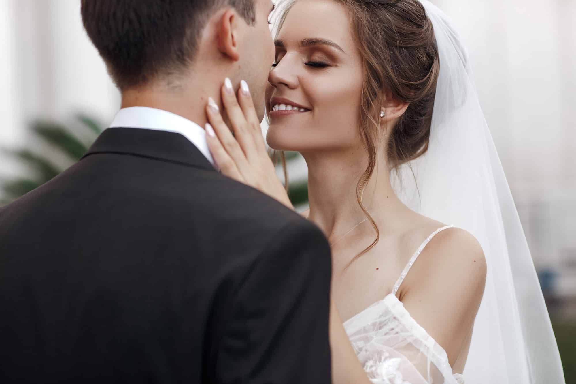Eine Braut in einem weißen Kleid und Schleier lächelt mit geschlossenen Augen und berührt zärtlich von hinten das Gesicht eines Mannes im schwarzen Anzug und fängt einen intimen Moment während ihrer Hochzeit ein.