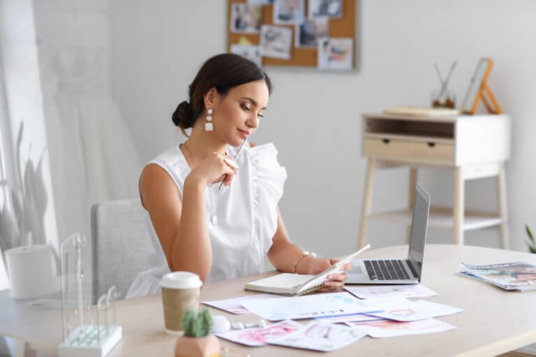 Eine Frau in einem weißen ärmellosen Top sitzt nachdenklich mit einem Notizbuch und einem Stift an einem Schreibtisch, umgeben von Hochzeitsmagazinen und ihrem Laptop. Eine Kaffeetasse und eine kleine Pflanze vervollständigen die Szene, während das schwarze Brett hinter ihr mit Fotos von den letzten Hochzeitsplanungssitzungen geschmückt ist.