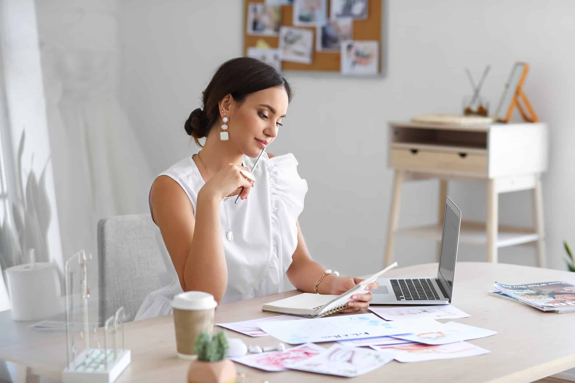 Eine Frau in einem weißen ärmellosen Top sitzt nachdenklich mit einem Notizbuch und einem Stift an einem Schreibtisch, umgeben von Hochzeitsmagazinen und ihrem Laptop. Eine Kaffeetasse und eine kleine Pflanze vervollständigen die Szene, während das schwarze Brett hinter ihr mit Fotos von den letzten Hochzeitsplanungssitzungen geschmückt ist.