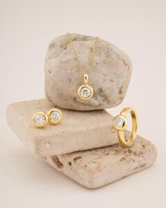Dieses exquisite Goldschmuckset ist perfekt für eine Hochzeit und besteht aus einer Halskette, einem Ring und Ohrringen mit runden Diamanteinsätzen. Elegant präsentiert auf strukturierten beigen Steinen vor einem neutralen Hintergrund verleihen diese Stücke jedem Brautensemble Raffinesse.