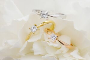 Drei Ringe mit Diamanten im Quadratschliff – einer aus Silber, einer aus Gold und einer aus Roségold – sind elegant auf weißen Blütenblättern angeordnet und fangen die Essenz von „heiraten“ ein.