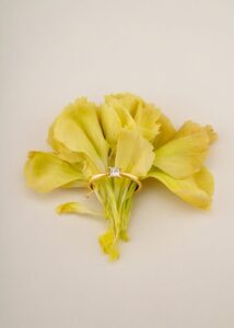 Ein goldener Ring mit einem Diamanten im Quadratschliff ist elegant auf einem Strauß zarter gelber Blumen vor einem neutralen Hintergrund platziert und fängt in seiner zeitlosen Schönheit die Essenz der Hochzeit ein.