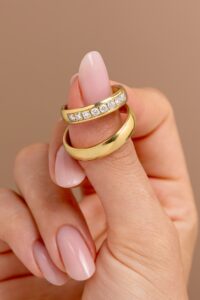 Eine Hand mit manikürten Nägeln hält zwei goldene Ringe, perfekte Symbole für eine Hochzeit. Ein Ring ist mit einer Reihe kleiner Diamanten verziert, die Ihrer Hochzeitsplanung Glanz verleihen, während der andere schlichtes Gold ist. Der Hintergrund ist in einem sanften Beige gehalten.