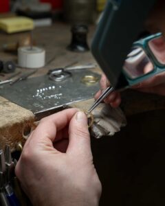 Eine Person verwendet ein Vergrößerungsstirnband, um einen Ring an der Werkbank eines Juweliers zu untersuchen und zu bearbeiten, was für alle, die eine Hochzeit planen, unverzichtbar ist. Im Hintergrund sind verschiedene Werkzeuge und Materialien verstreut, die alle eine Rolle bei der Herstellung des perfekten Symbols des Heiratens spielen.