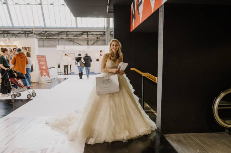 Eine Frau im Brautkleid steht mitten im Trubel der Hochzeitsplanung drinnen und hält eine Einkaufstasche und einige Papiere in der Hand. Im Hintergrund sind Menschen und Schaufenster zu sehen, die Umgebung ist gut beleuchtet und die Architektur modern.