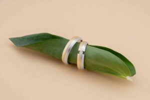 Zwei goldene Ringe ruhen auf einem grünen Blatt vor einem beigen Hintergrund. Ein Ring ist mit einem kleinen Diamanten verziert, während der andere schlicht ist. Die Komposition ist schlicht und elegant, perfekt für jede Hochzeit und betont den Kontrast zwischen dem metallischen Glanz und der natürlichen Textur.