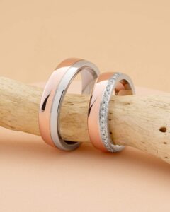 Zwei elegante Ringe auf einem Stück Treibholz erinnern an die Essenz der Hochzeitsplanung. Einer ist ein einfacher Ring aus Roségold und Silber, während der andere ein Roségoldring ist, der mit funkelnden Diamanten auf einem Silberstreifen verziert ist, perfekt für diejenigen, die sich auf ihre Hochzeit vorbereiten. Vor einem weichen beigen Hintergrund.