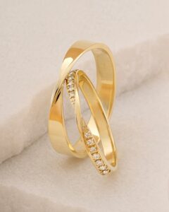 Zwei ineinander verschlungene goldene Eheringe auf einer weichen, cremefarbenen Oberfläche fangen die Essenz einer Hochzeit ein. Ein Ring ist schlichtes Gold, während der andere auf der Hälfte seines Bandes mit kleinen Diamanten verziert ist, was ein Gefühl von Eleganz und Luxus vermittelt, das perfekt für jede Hochzeitsplanung ist.