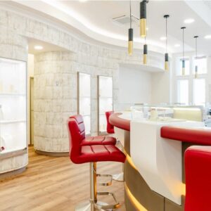 Modernes Salon-Interieur mit geschwungenen weißen Wänden und Holzboden. Rote Lederbarhocker stehen um eine weiß-rote Theke herum und machen es zu einem idealen Ort für Hochzeitsplanung. Elegante Beleuchtungskörper hängen von der Decke und schaffen eine helle und einladende Atmosphäre für Ihren besonderen Tag.