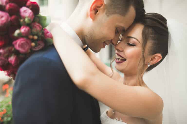 An ihrem Hochzeitstag umarmen sich Braut und Bräutigam freudig. Die Braut trägt einen Schleier und lächelt breit, während sie einen Strauß rosa Blumen in der Hand hält. Der Bräutigam in einem marineblauen Anzug sieht sie mit einem glücklichen Gesichtsausdruck an, während sie diesen wunderschönen Moment der Hochzeit feiern.