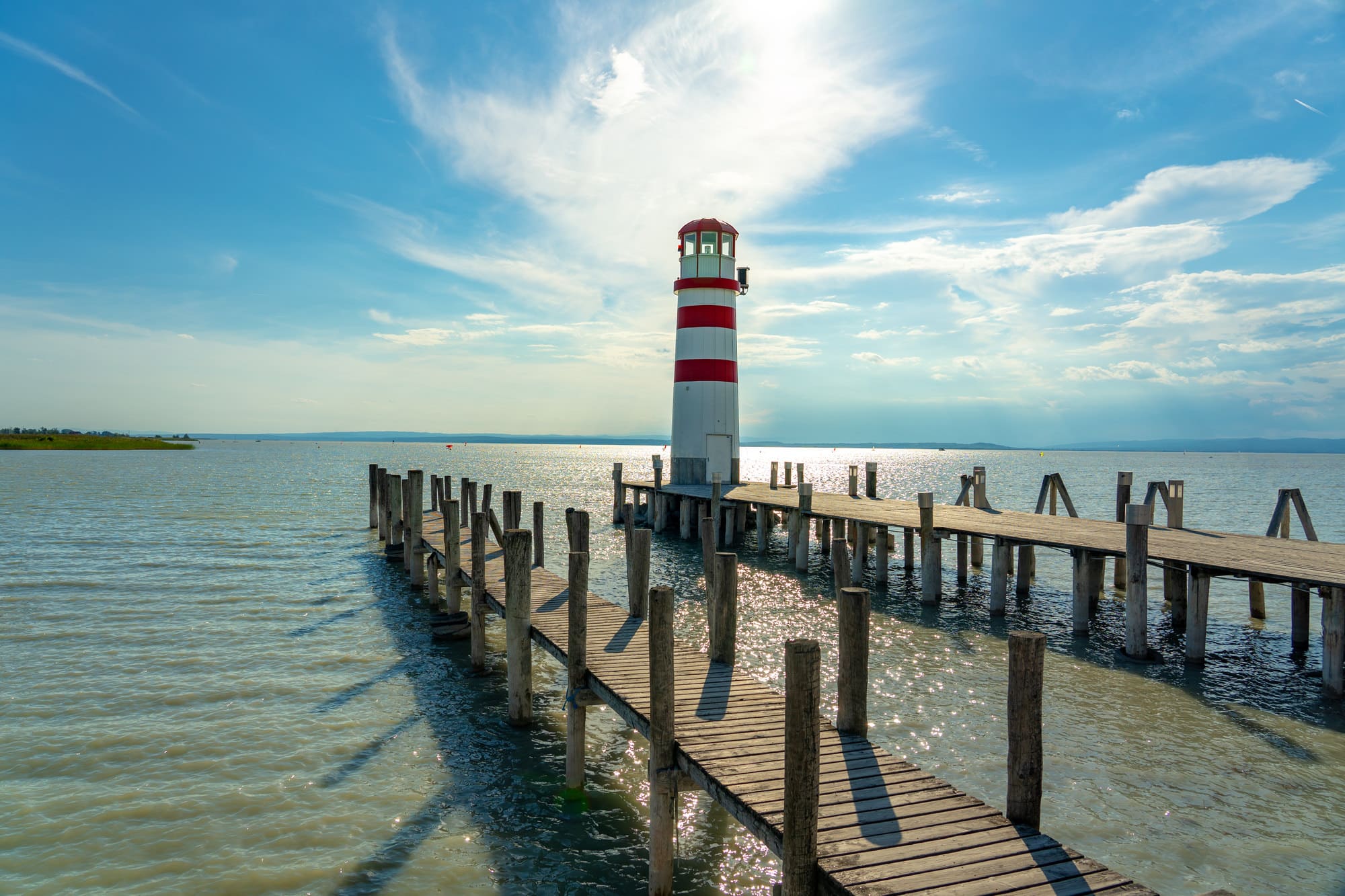Ein Holzsteg führt zu einem rot-weiß gestreiften Leuchtturm, der in ruhigem Wasser unter einem blauen Himmel mit vereinzelten Wolken steht. Das Sonnenlicht spiegelt sich im Wasser und schafft eine friedliche und heitere Atmosphäre – perfekt für eine romantische Hochzeit oder eine intime Hochzeitsplanung.