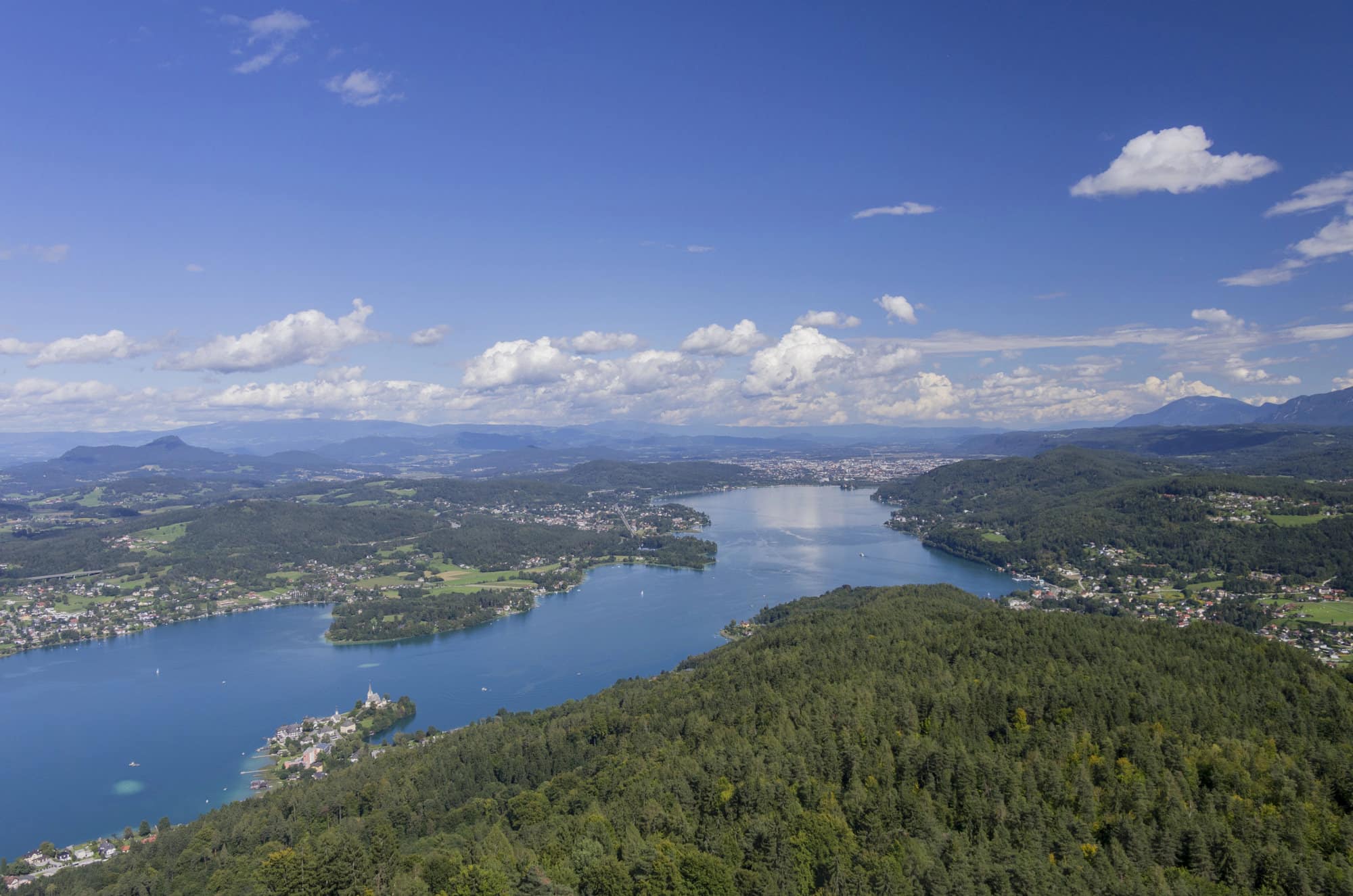 Eine malerische Luftaufnahme eines großen Sees, umgeben von üppigen grünen Wäldern und sanften Hügeln unter einem klaren blauen Himmel, bietet die perfekte Kulisse für eine Traumhochzeit. Kleine Städte und vereinzelte Wolken ergänzen die malerische Landschaft und schaffen eine ruhige Atmosphäre, die ideal für romantische Gelübde ist.