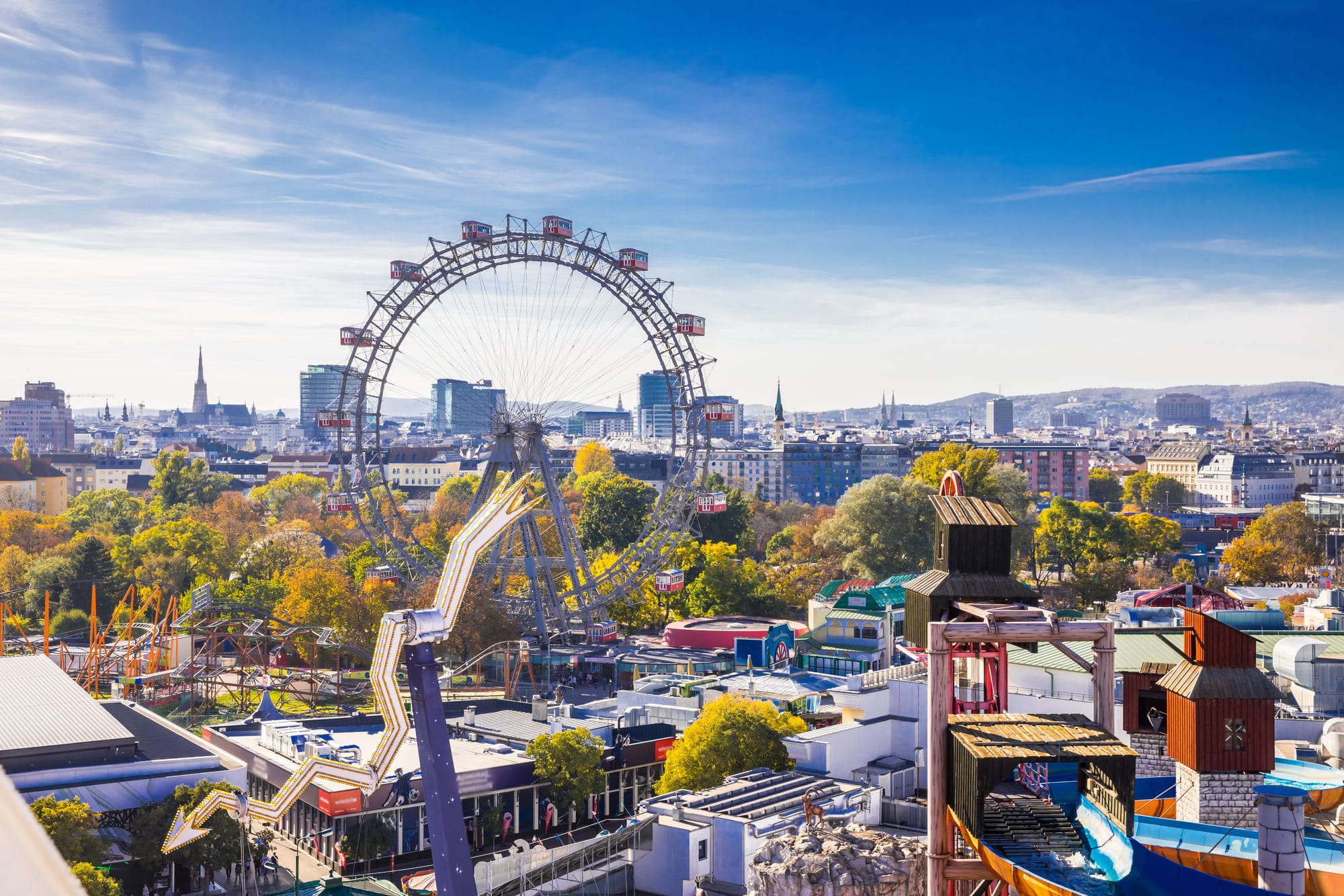 Eine lebendige Stadtlandschaft mit einem Vergnügungspark und einem markanten Riesenrad – perfekt als einzigartige Hochzeitskulisse. Bunte Buden und Fahrgeschäfte verschmelzen nahtlos mit städtischen Gebäuden unter einem klaren blauen Himmel und fangen die Essenz lebhafter Feiern an einem sonnigen Tag ein.