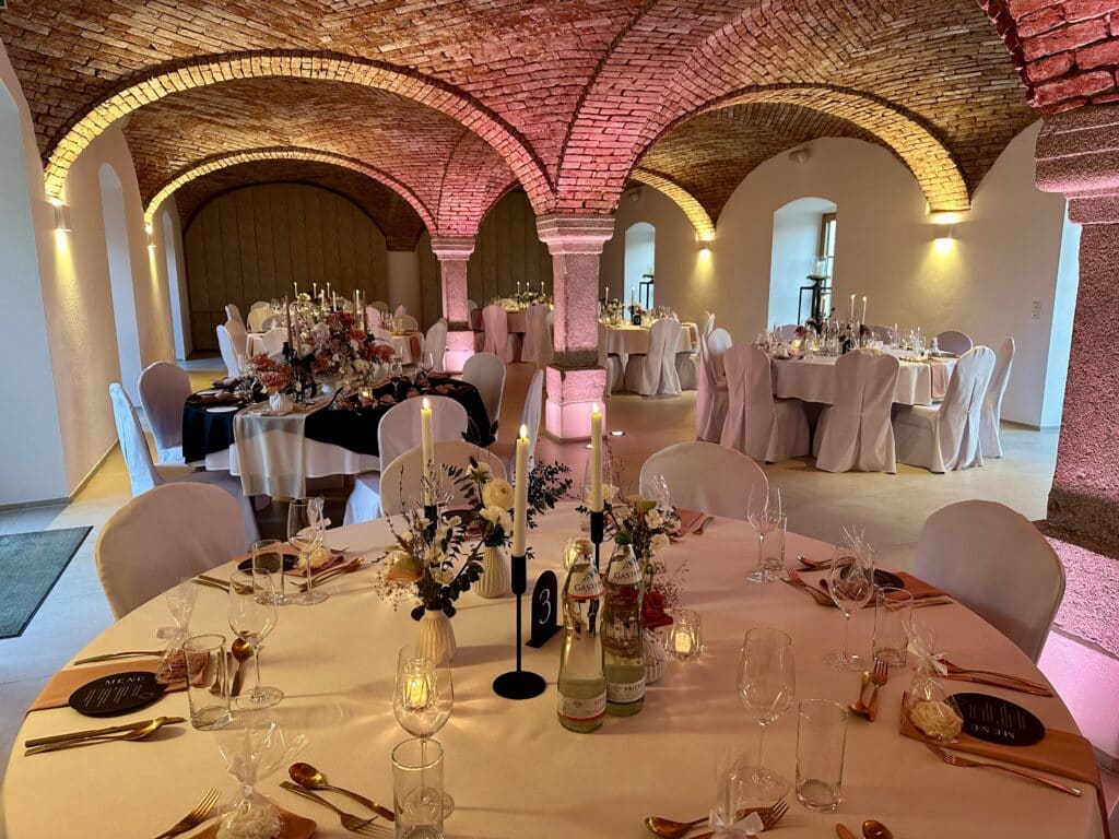 Ein wunderschön dekorierter Veranstaltungsort für Hochzeitsempfänge mit runden Tischen für die Gäste. Jeder Tisch ist mit eleganten Blumendekorationen, Kerzen und Gedecken ausgestattet. Der Raum hat Backsteinbögen und sanftes Umgebungslicht, was eine warme und einladende Atmosphäre schafft, die perfekt zum Heiraten ist.