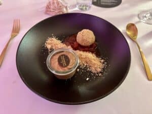 Ein Gourmet-Dessert, das sich ideal für eine Hochzeit eignet, wird auf einem schwarzen Teller präsentiert und besteht aus einem runden, mit Krümeln überzogenen Leckerbissen, einer Kugel Sorbet auf Fruchtkompott und einem kleinen Glas mit cremiger Schokoladenmousse, gekrönt mit einer Schokoladenmünze. Elegantes goldenes Besteck begleitet das Gericht.