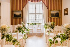 Ein wunderschön dekorierter Raum für eine Hochzeitszeremonie verfügt über weiße Stühle, die mit Grünpflanzen und Blumen geschmückt sind und in Reihen vor einem großen Fenster mit gestreiften Vorhängen aufgestellt sind. Gerahmte Gemälde schmücken die Wände und verleihen eine elegante Note, ideal für diejenigen, die gerade mit der Hochzeitsplanung beschäftigt sind.