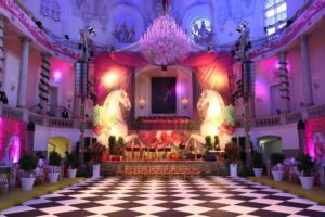 Ein großer Ballsaal mit einem großen Kronleuchter, thematischen Dekorationen mit Pferden und einem farbenfrohen Hintergrund, der sich perfekt für die Hochzeitsplanung eignet. Der schwarz-weiß karierte Boden ergänzt die für die Veranstaltung aufgestellten Stuhlreihen. Die rosa-violette Beleuchtung verleiht Ihrer Hochzeit eine romantische Note.