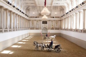 Eine große Indoor-Arena mit kunstvollen Kronleuchtern und Säulen, ideal für eine königliche Hochzeit. Zwei Pferdekutschen, in denen jeweils eine Person sitzt, gleiten anmutig über den Sandboden. Die Kulisse verbindet Eleganz mit Geschichte und ist perfekt für alle, die von einer märchenhaften Hochzeit träumen.