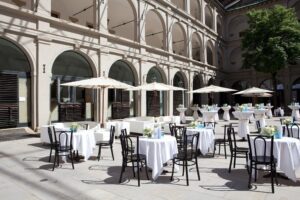 Ein Café im Freien, perfekt für eine Traumhochzeit, mit runden Tischen mit weißen Tischdecken und schwarzen Stühlen unter Sonnenschirmen an einem sonnigen Tag. In einem Innenhof mit hohen Bögen und einem Schatten spendenden Baum gelegen, bietet es eine elegante Atmosphäre, ideal für jede Hochzeitsplanung.