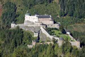 Luftaufnahme eines großen, historischen Schlosses am Hang, das sich perfekt für die Hochzeitsplanung eignet und von dichtem grünem Wald umgeben ist. Das Schloss verfügt über mehrere Türme und ein zentrales Hauptgebäude mit einer grünen Kuppel. Eine Steinmauer umschließt das Gelände, das einen Weg und Gärten umfasst.