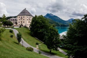 Ein malerischer Blick auf ein historisches Gebäude, umgeben von üppigem Grün, perfekt für eine Traumhochzeit, mit Blick auf einen ruhigen blauen See. Im Hintergrund erheben sich bewaldete Hügel und ein Berg unter einem wolkigen Himmel. Ein gewundener Pfad führt durch diese malerische Landschaft, ideal für romantische Heiratsmomente.