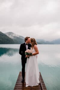 Braut und Bräutigam küssen sich während ihrer Hochzeit auf einem Holzsteg an einem ruhigen, nebligen See. Die Braut hält einen Strauß rosa und weißer Blumen in der Hand, im Hintergrund sind Berge unter einem bewölkten Himmel zu sehen.